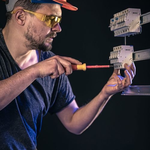 A male electrician in a protective helmet works in a switchboard with an electrical connecting cable.